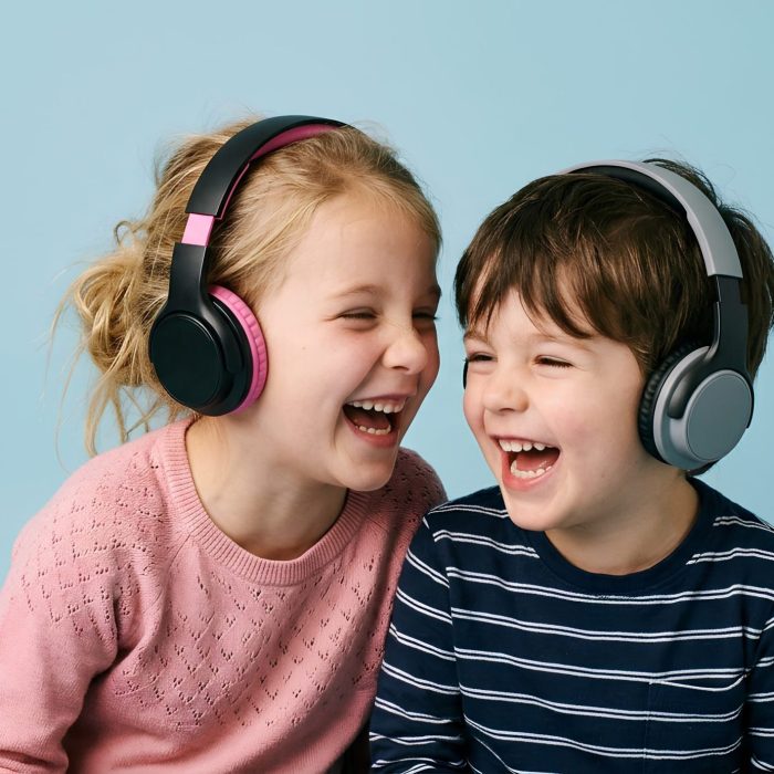 Cheerful young girl and her brother wearing over-ear headphones against a seamless soft-blue backdrop, both laughing joyfully together in studio portrait